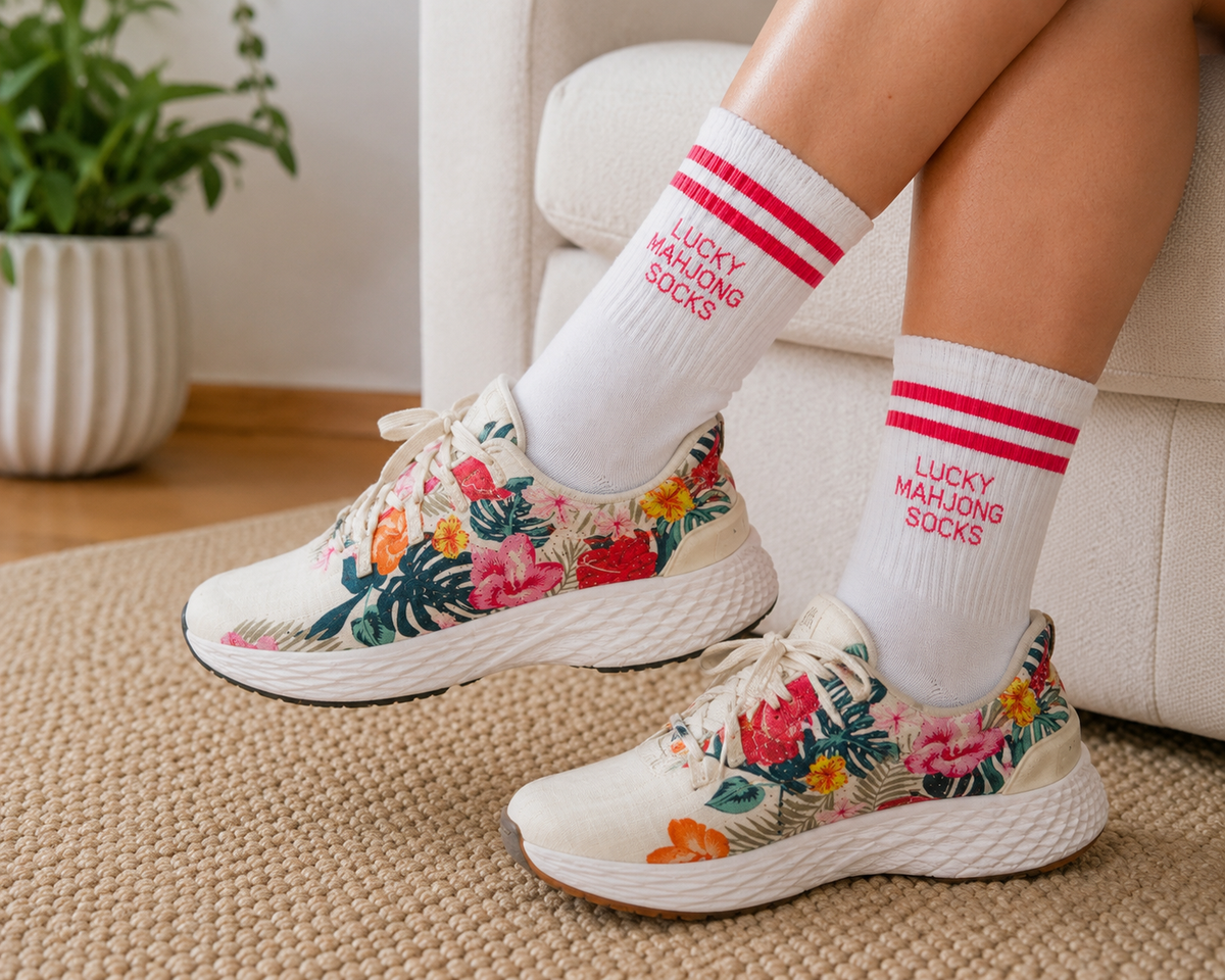 A person seated indoors wearing white crew socks with red stripes and the text 'LUCKY MAHJONG SOCKS' paired with white sneakers featuring a colorful tropical floral print. 